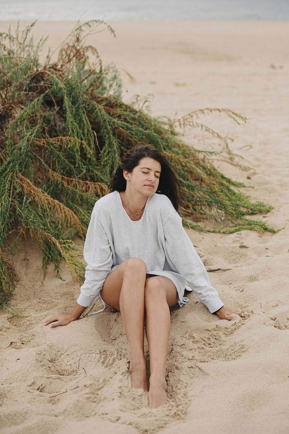 A woman in a grey sweatshirt sitting on sand.